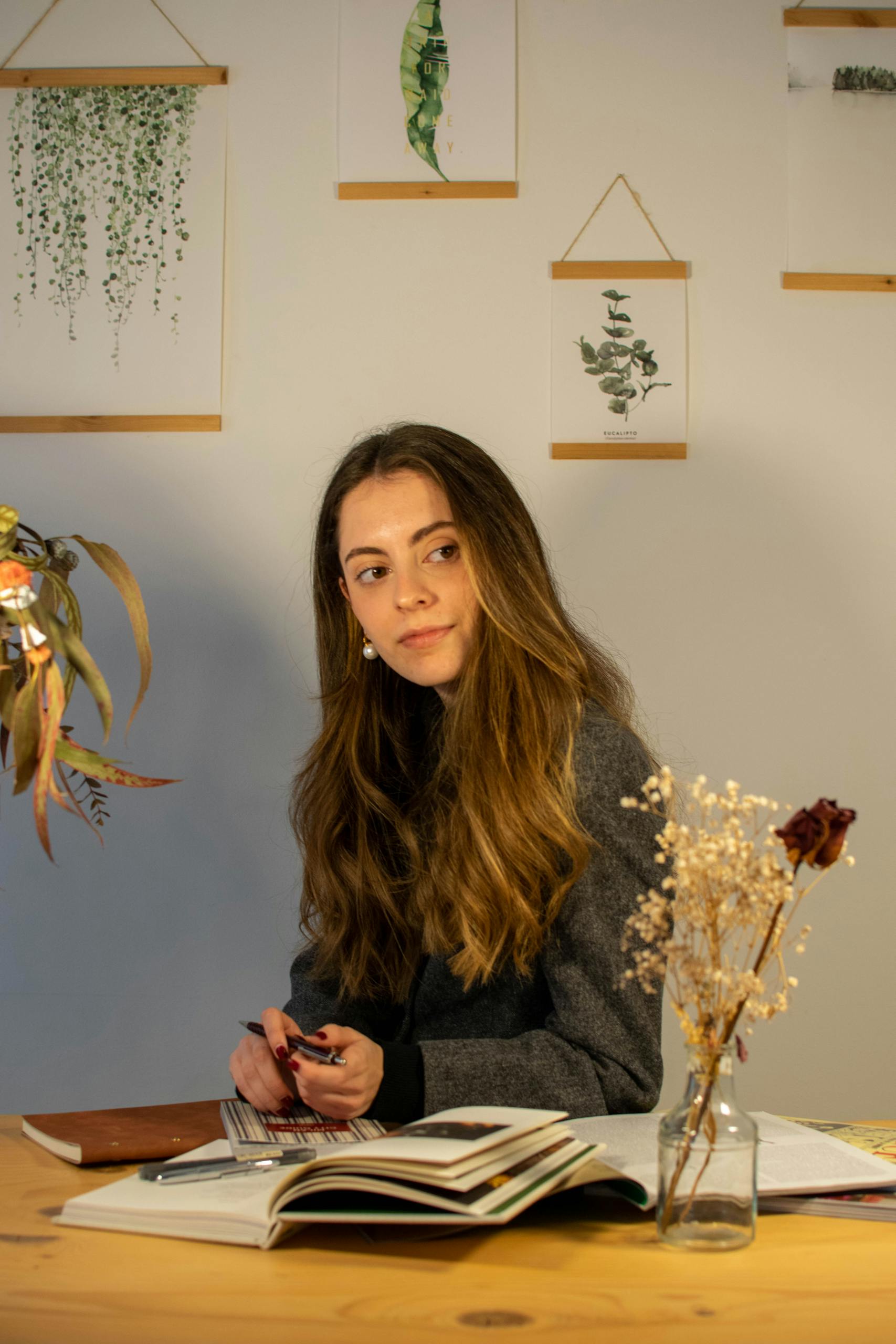A young woman sits at a desk writing, surrounded by books and plants, creating a cozy work atmosphere.